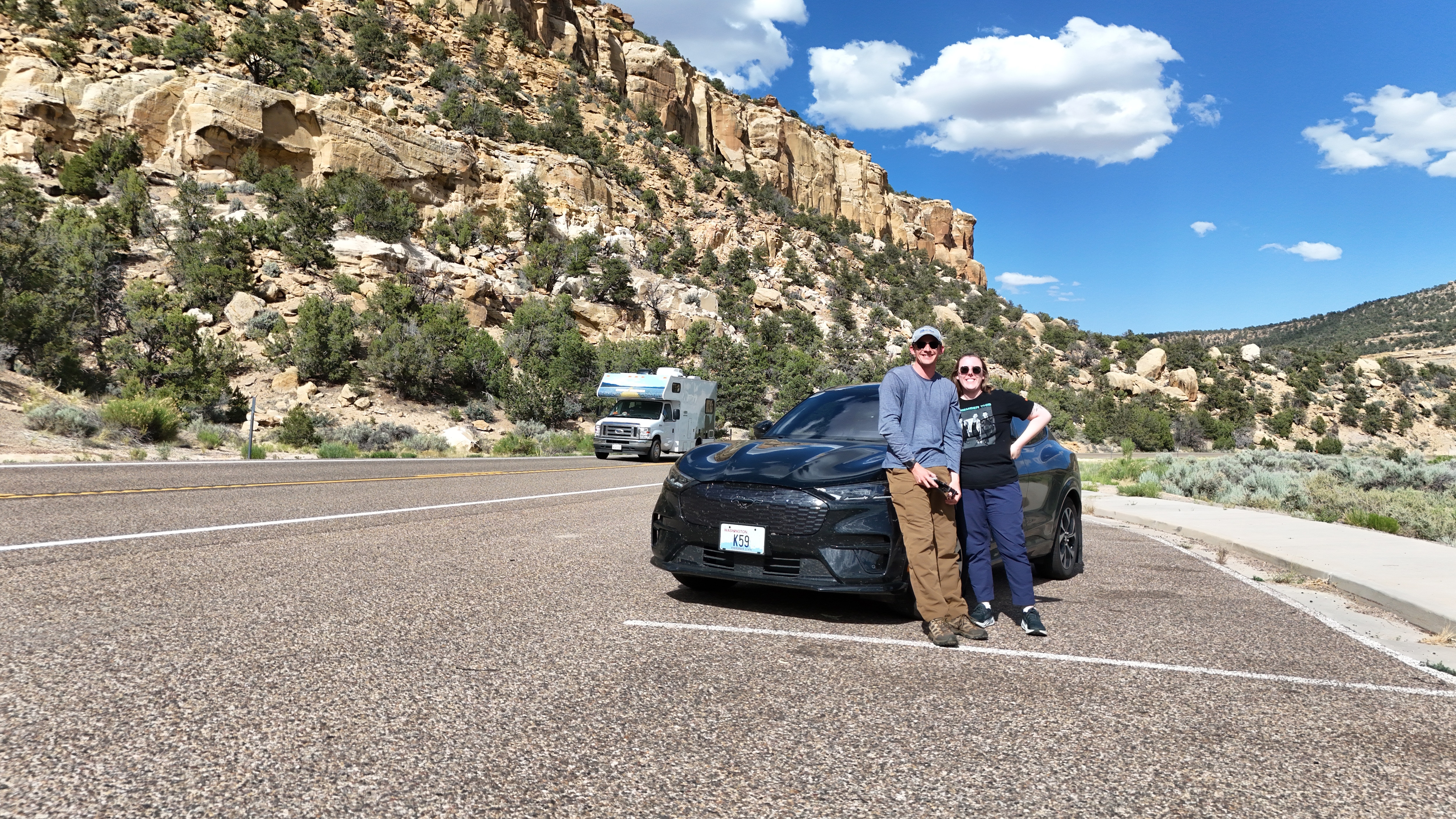 A couple poses near a Ford Mustang Mach-E next to a scenic drive.